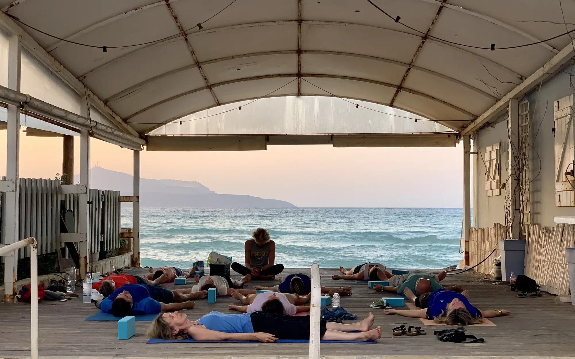 An evening yoga class lying in relaxation pose under a canopy beside the sea