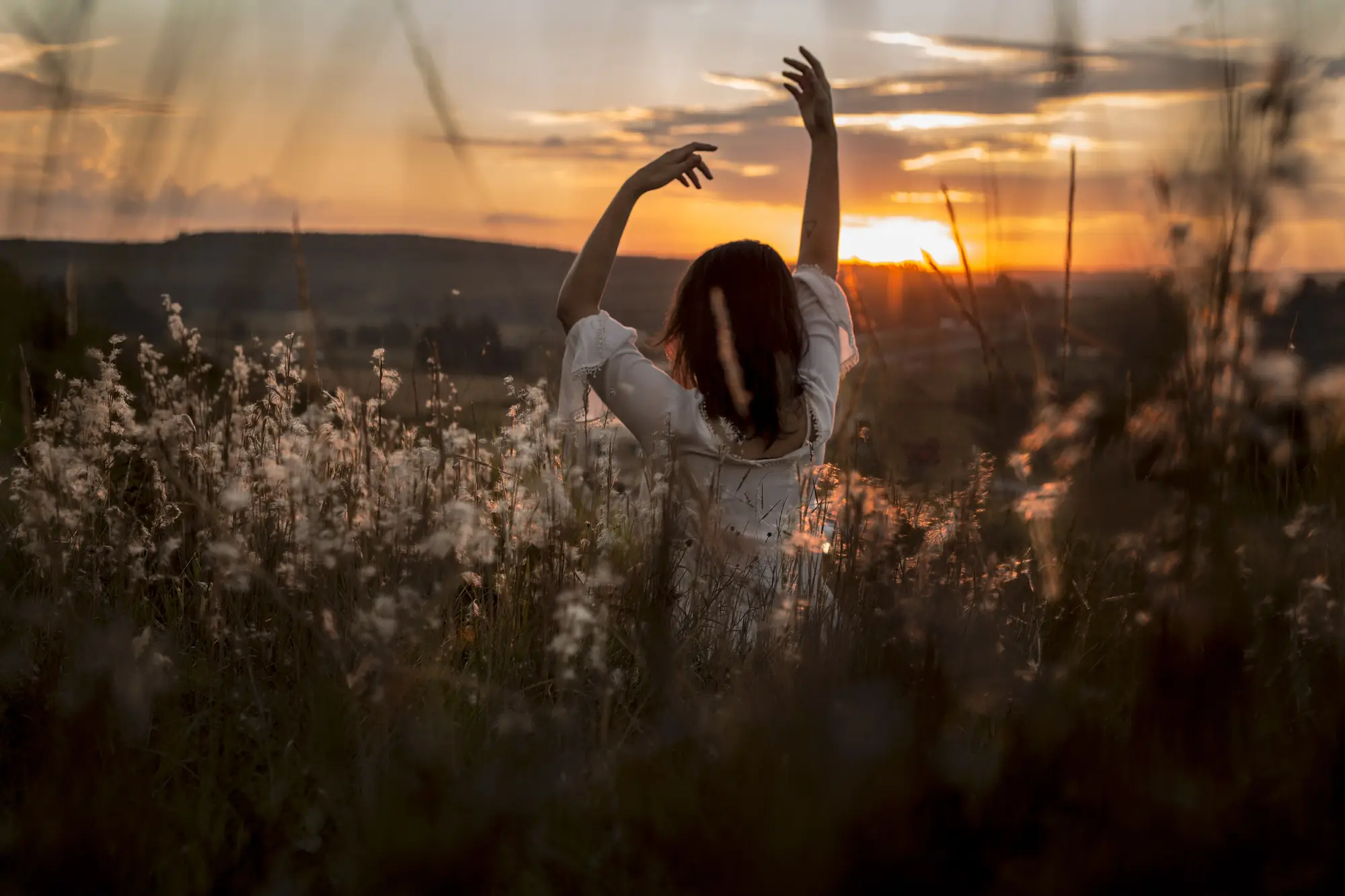 A woman raising her arms overhead in a gentle standing stretch at sunset