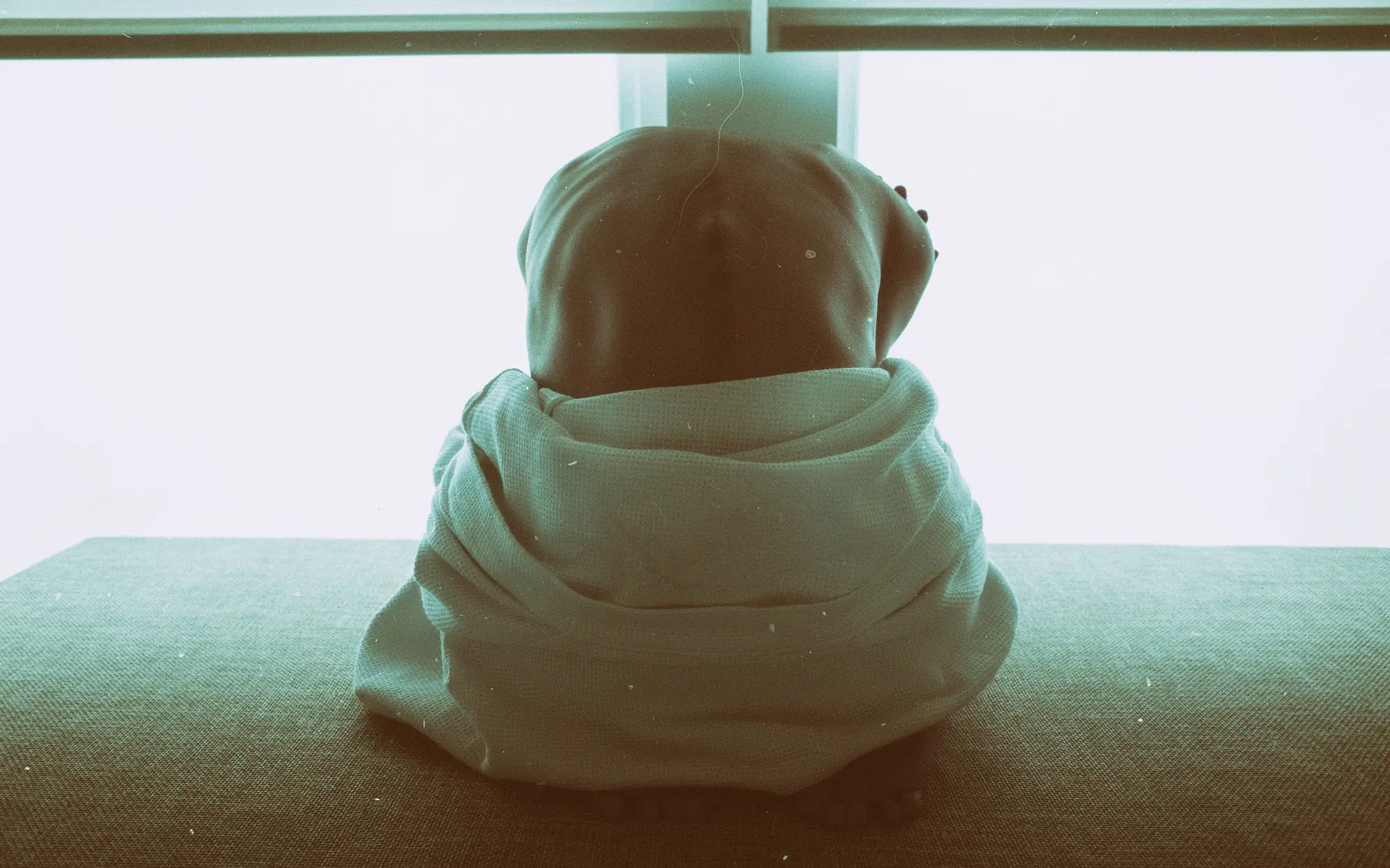 A person wrapped in a hooded sweater, sitting quietly on a mat facing a window