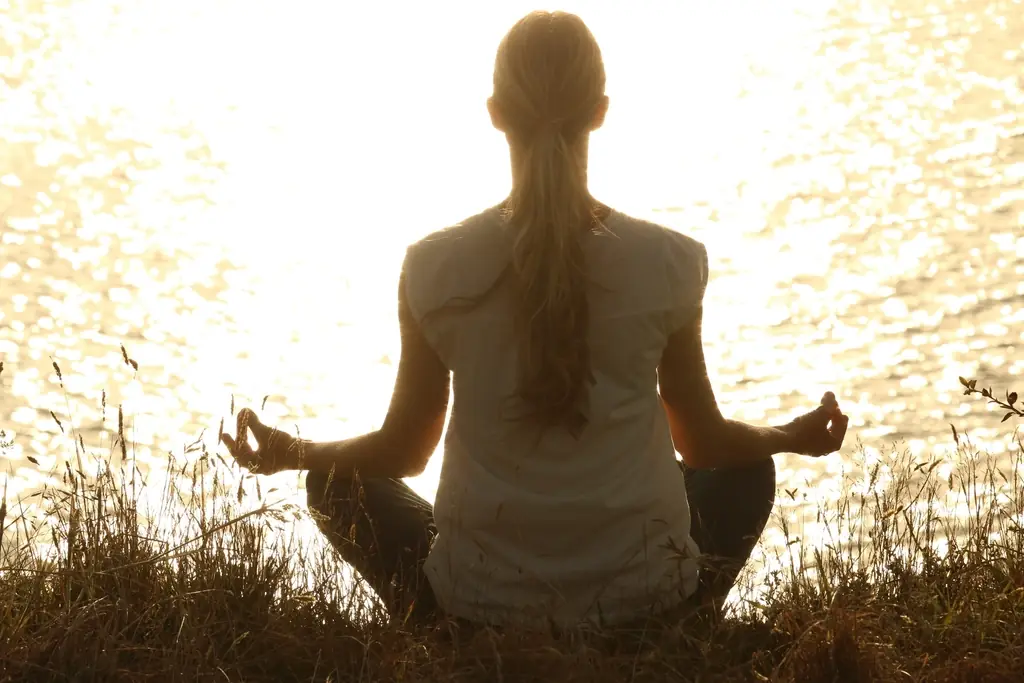 A woman sitting cross-legged with her hands resting on her knees, facing sunlit water