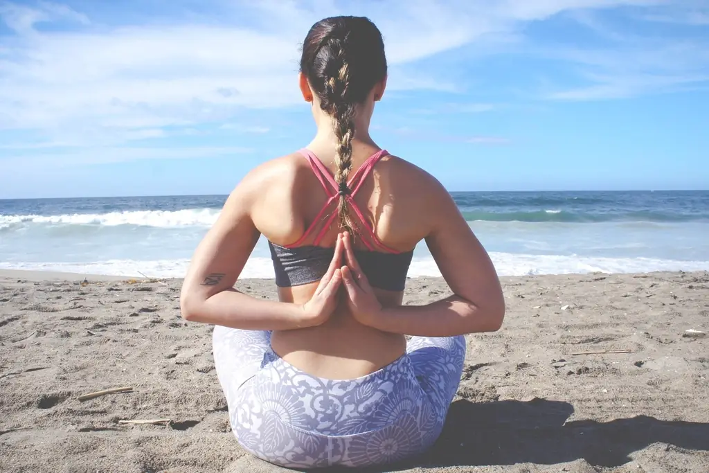 A woman sitting cross-legged on the sand with an upright spine, facing the sea