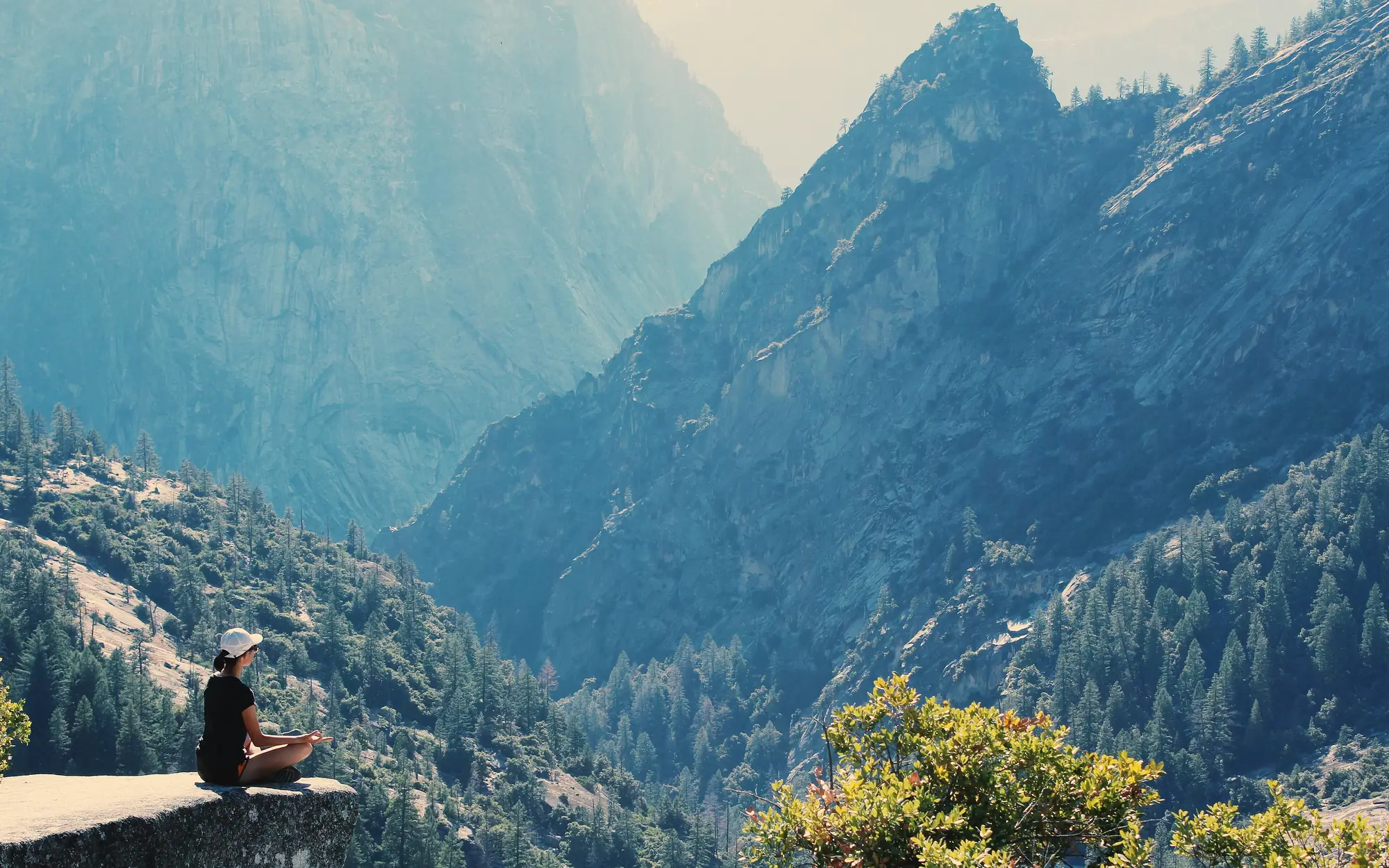 A person sitting cross-legged on a rock ledge above a mountain valley, practising breathing
