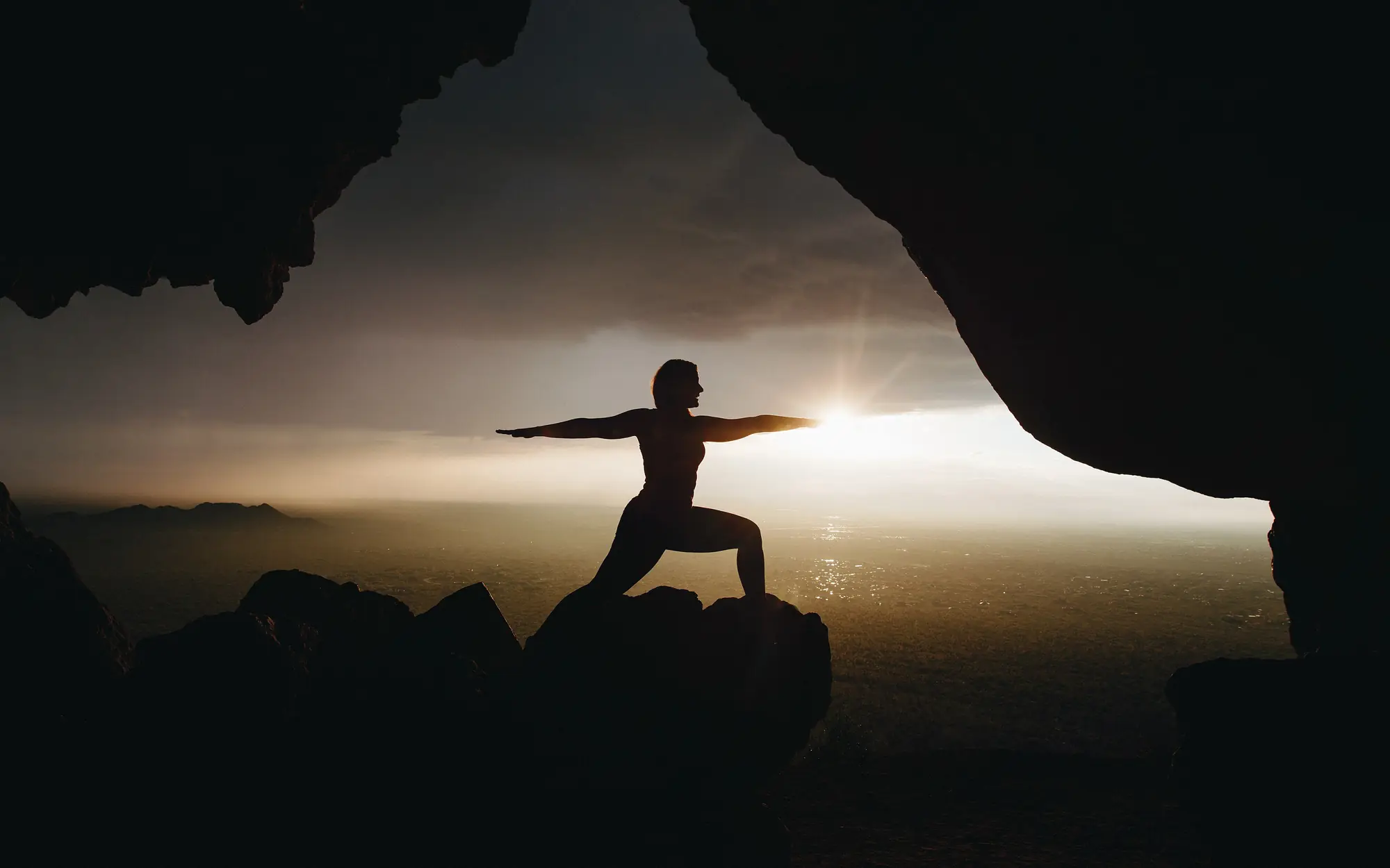 Silhouette of a woman holding Warrior II pose on a rock at sunrise